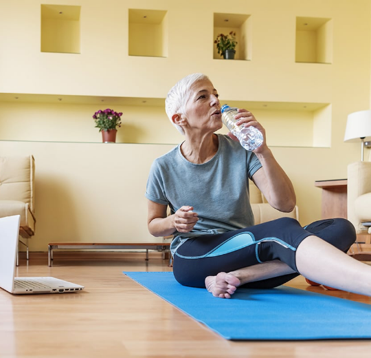 Picture: Woman sitting on yoga mat inside drinking out of water bottle with open laptop next to her