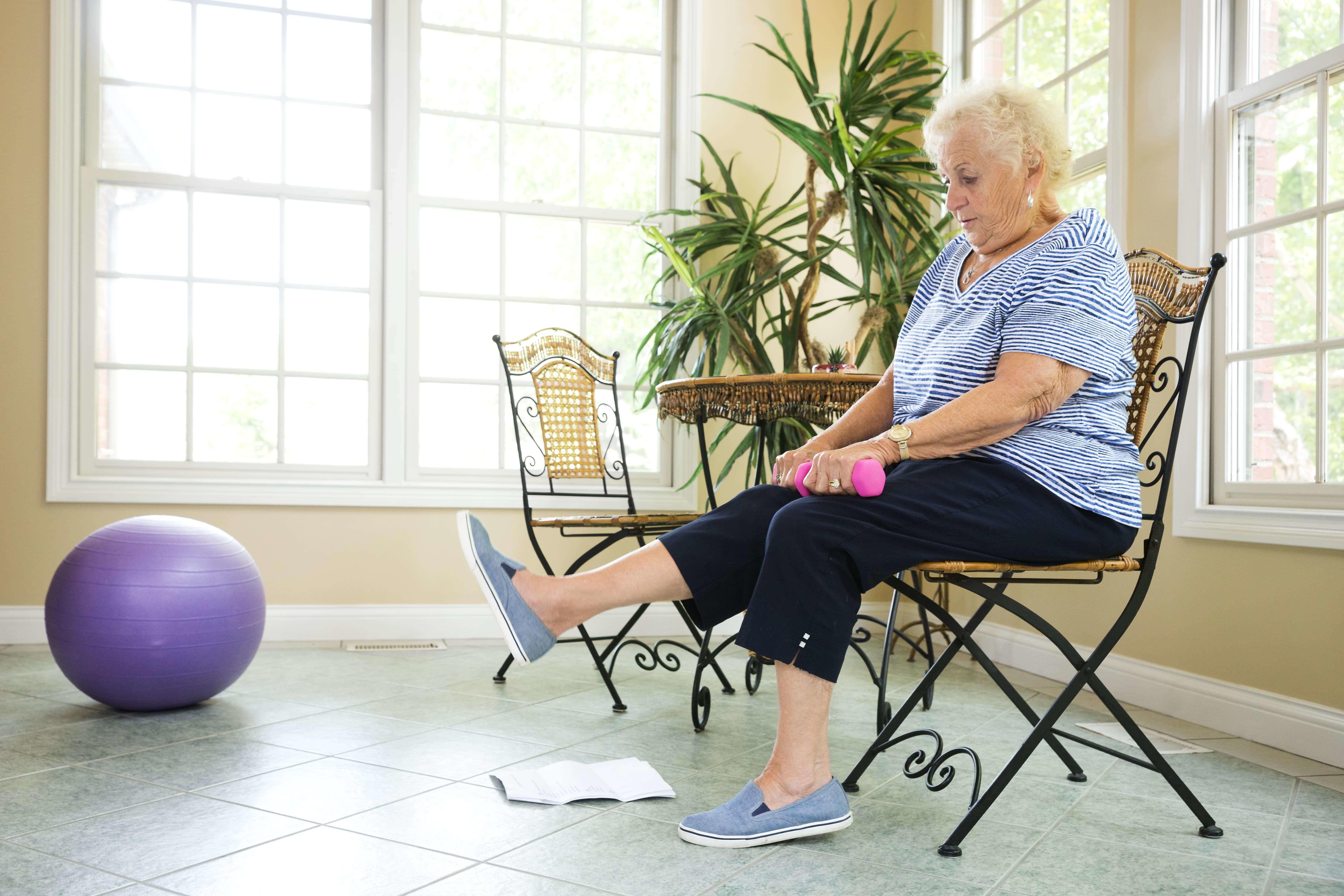 Woman sits in chair, holds light weights and raises her leg while performing exercises at home