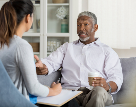 Senior man sitting on couch facing talking to female therapist taking notes