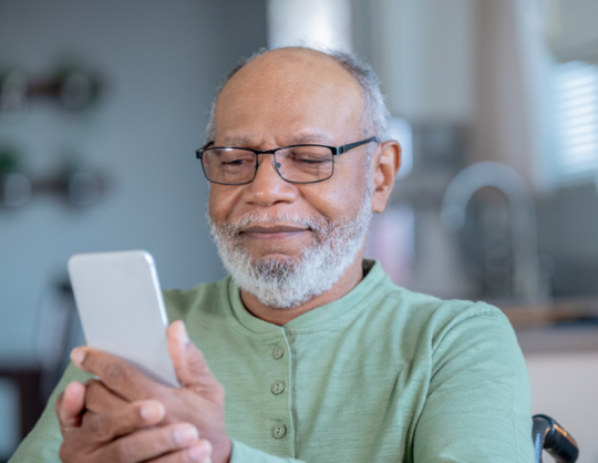 Man with glasses sits at kitchen table while looking at cell phone in hands