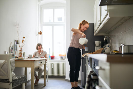 Woman sitting at kitchen table smiling while caregiver stands and pours coffee into mug