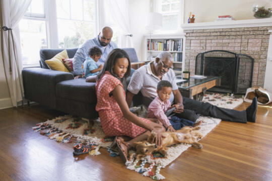 Mature man sitting on floor with granddaughter and grandson petting dog while adult son sits on couch with other young child