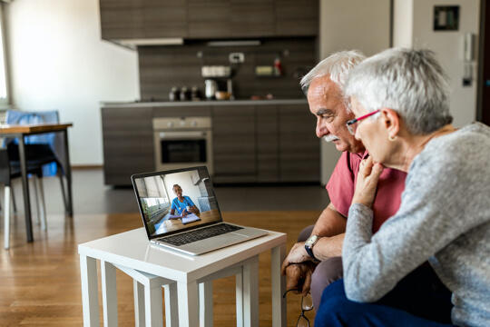 Woman wearing glasses sitting next to man holding glasses while looking at laptop computer with doctor speaking on screen