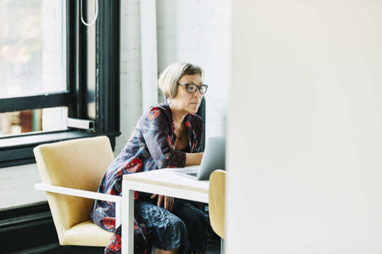 Woman with glasses sitting in chair at desk looking at laptop