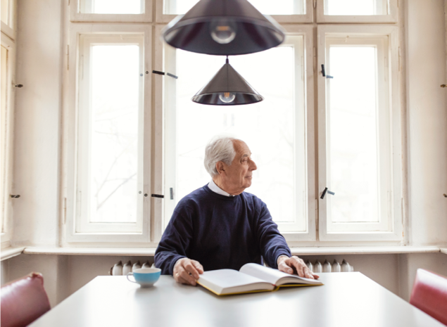 Older man reading book at kitchen table underneath lighting fixtures
