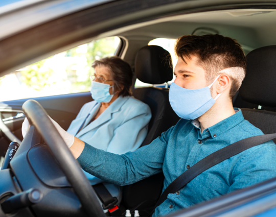 Man wearing a face mask driving with an older woman wearing glasses and a face mask sitting in passenger seat